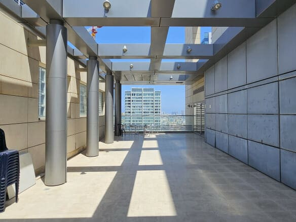 שביל חיצוני מקורה ברחוב רוטשילד עם עמודי מתכת ולוחות זכוכית בגג; כיסאות מוערמים נמצאים משמאל, ובניינים גבוהים נראים ברקע.