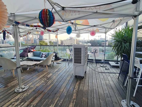 מרפסת גג חיצונית עם רצפת עץ, פינת ישיבה, כסאות בר, מזגן נייד וקישוטים צבעוניים מנייר תחת סככה. משקיפה על קו הרקיע של העיר וקרובה לתחנת רכבת השלום — מושלמת לכל מפגש משרדי.