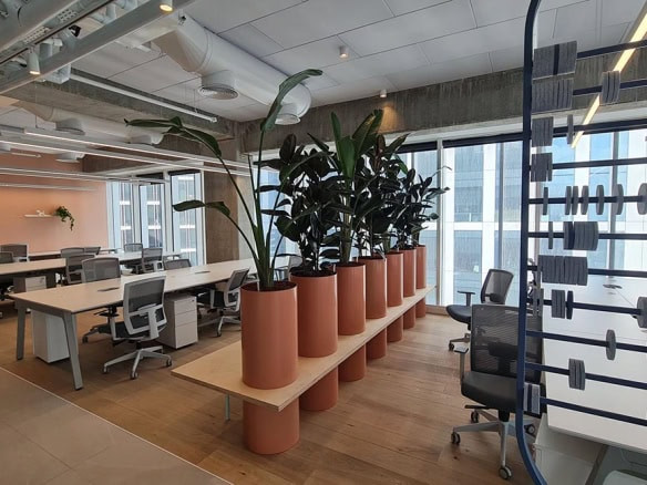 משרד מודרני בעל חלל פתוח עם שולחנות, כיסאות, עציצים גדולים על ספסל וחשבונייה דקורטיבית ליד חלונות גבוהים המשקיפים על נוף העיר של מגדל סקיי – מושלם עבור השכרת משרדים.
