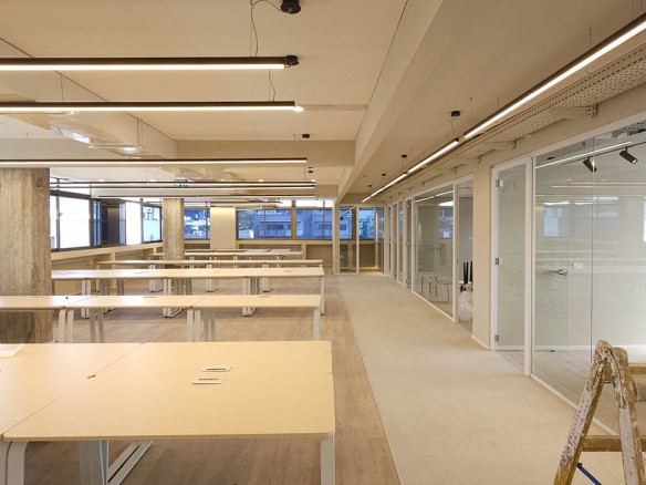 משרד להשכרה מודרני בעל חלל פתוח בבנייה ברחוב מנחם בגין, עם שולחנות ריקים, מחיצות זכוכית, תאורת תקרה חשופה וסולם עץ בחזית.