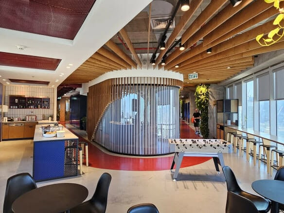 אזור מנוחה מודרני להשכרה במידטאון, הכולל שולחנות עגולים, שולחן כדורגל, עמדת קפה, קורות עץ בתקרה וחלונות גדולים עם נוף לעיר וצמחייה עשירה בקומה עצמאית.