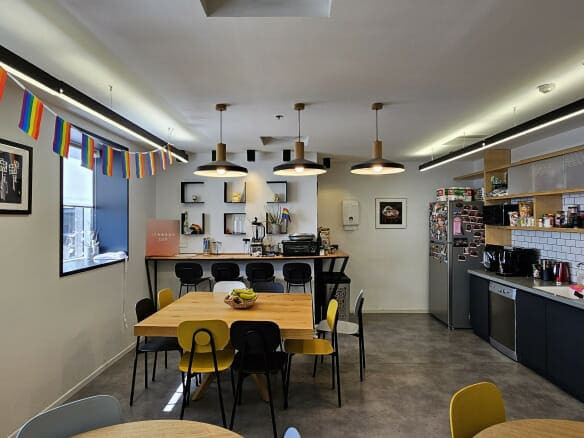 מטבח משרדי מודרני במגדל מבוקש, הכולל שולחנות, כיסאות, מכשירי חשמל, מדפים פתוחים ודגלי גאווה. החלל המואר כולל מכונת קפה ואספקה – אידיאלי למי שמחפש משרד להשכרה עם מתקנים מעולים.