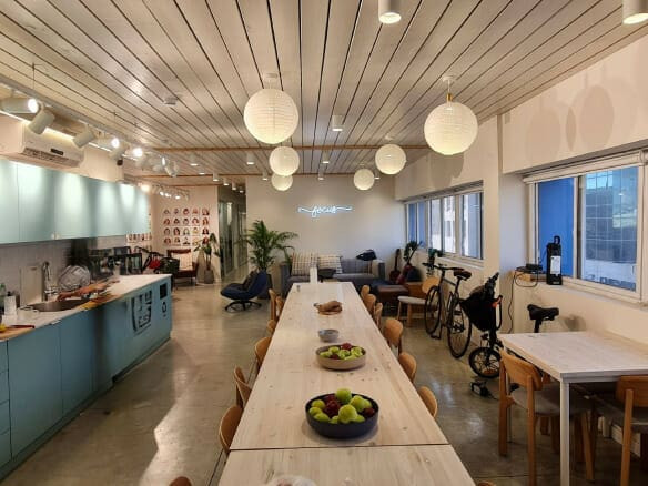 מטבח משותף מודרני ופינת אוכל עם שולחנות ארוכים, קערות פירות, ארונות בצבע תכלת, פנסי נייר תלויים וחלונות גדולים בצד ימין — אידיאלי עבור משרד להשכרה בשכונת מונטיפיורי, תל אביב.