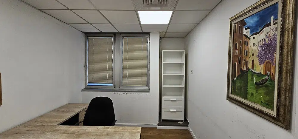 משרד קטן להשכרה במגדל רובינשטיין, הכולל שולחן עץ, כיסא שחור, שני חלונות עם תריסים, מדף לבן ותמונה ממוסגרת על הקיר. שטח המשרד 60 מ
