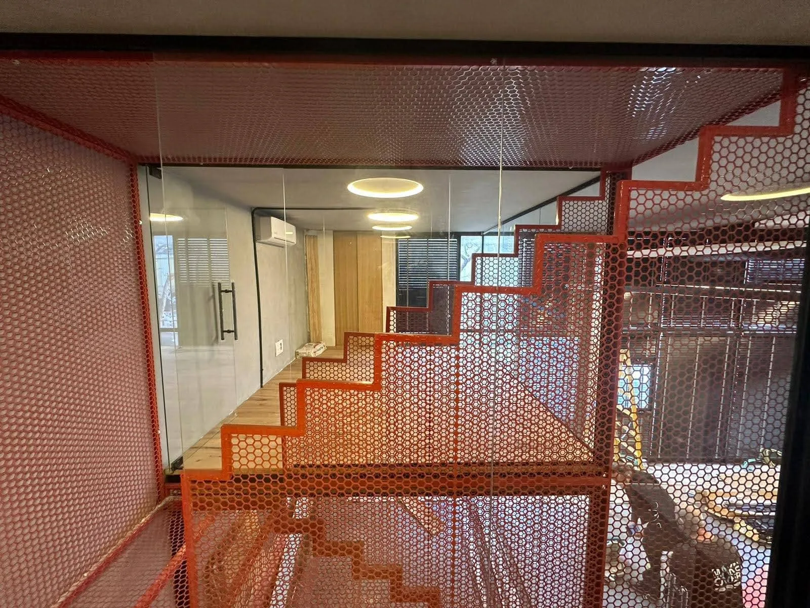 מדרגות מתכת אדומות בעיצוב זוויתי בתוך חלל פנים מודרני בסגנון תעשייתי, עם קירות זכוכית, נגיעות עץ ותאורת תקרה עגולה.
