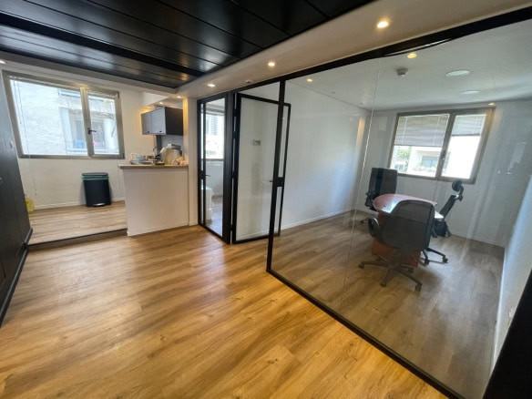 חלל משרדי מודרני עם ריצוף עץ, חדר ישיבות עם קירות זכוכית, שולחן, כיסאות ומטבחון, הכל מואר בתאורת תקרה ובאור טבעי מהחלונות.