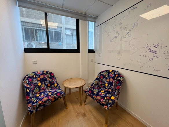 שני כיסאות מעוצבים ושולחן עגול קטן מסודרים בפינת המשרד, שעל קיריתו לוח לבן מכוסה בדיאגרמות והערות.