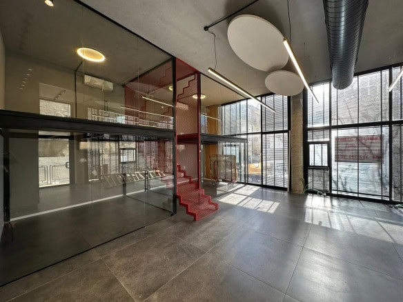 חלל משרדים מודרני וריק עם קירות זכוכית גדולים, תקרות גבוהות, תאורת תקרה עגולה ומדרגות אדומות מרכזיות עם דפנות מתכת. אור טבעי זורם מבעד לחלונות גבוהים.