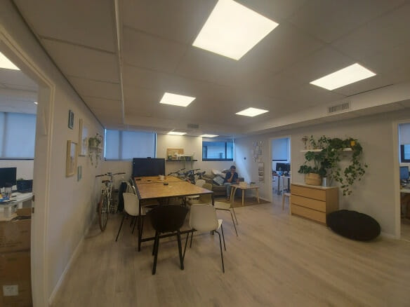 חלל משרדי מודרני עם שולחן מרכזי, כיסאות שונים, אופניים, צמחים, ובחורה יושבת על ספה ומשתמשת במחשב נייד ברקע.