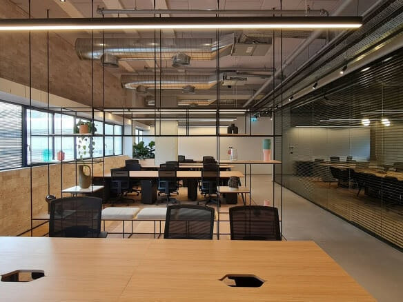 משרד מודרני בעל חלל פתוח עם שולחנות, כיסאות, חלונות גדולים, תעלות חשופות ומחיצות זכוכית המפרידות בין אזורי העבודה.