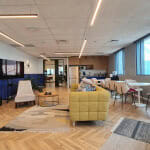 טרקלין משרדי מודרני עם ספה צהובה, כורסאות, טלוויזיה, שולחנות עגולים, כיסאות ועציצים, עם ריצוף עץ וחלונות גדולים המאפשרים כניסת אור טבעי.