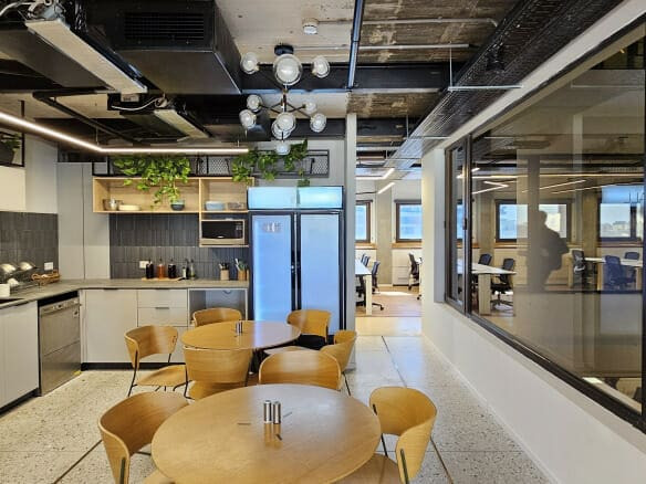 מטבח משרדי מודרני עם שולחנות עץ עגולים וכסאות, תאורה תקרתית, צמחים ונוף אל חלל עבודה סמוך מבעד לחלונות פנים גדולים — מושלם משרדים להשכרה בלב העיר בקומה עצמאית.