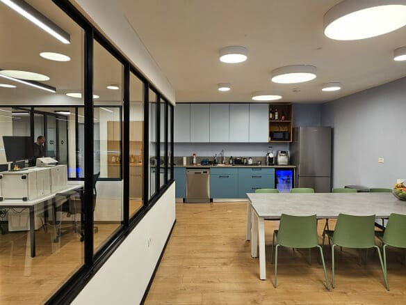 מטבח ומטבח מודרני עם כיסאות ירוקים, שולחן גדול, מכשירי חשמל וקיר זכוכית המפריד אותו מפינת העבודה; מושלם עבור אלה המחפשים משרד להשכרה בשרונה. אדם נראה עובד ברקע.