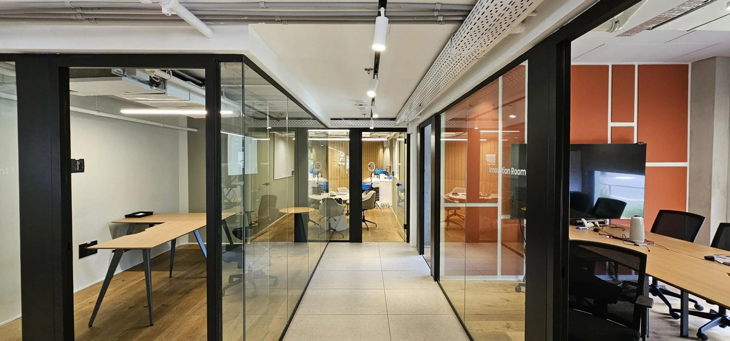 מסדרון משרדי מודרני עם חדרי ישיבות בעלי קירות זכוכית משני הצדדים, הכולל שולחנות עץ, כיסאות ותאורת תקרה.
