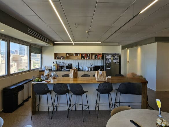 מטבח משרדי מודרני להשכרה במתחם שרונה, הכולל אי מעץ, כיסאות שחורים, שקיות נייר על הדלפק ונוף לעיר מבעד לחלונות גדולים. מדפים ומכשירים חשמליים מסודרים לאורך הקיר האחורי.