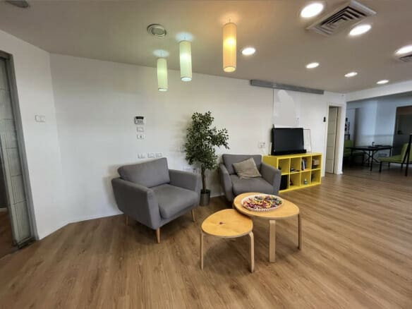 סלון מודרני עם שני כורסאות אפורות, שולחנות עץ קטנים, מדף קוביות צהוב עם טלוויזיה וקערת ממתקים על השולחן, המונח על רצפת עץ — אידיאלי עבור משרד ל-20 עובדים או כטרקלין מזמין במונטיפיורי.