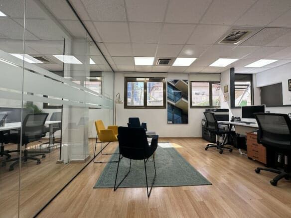 חלל משרדי מודרני עם קירות זכוכית, שולחנות, כיסאות משרדיים, שטיח ירוק ושני כורסאות צבעוניות ליד החלונות.