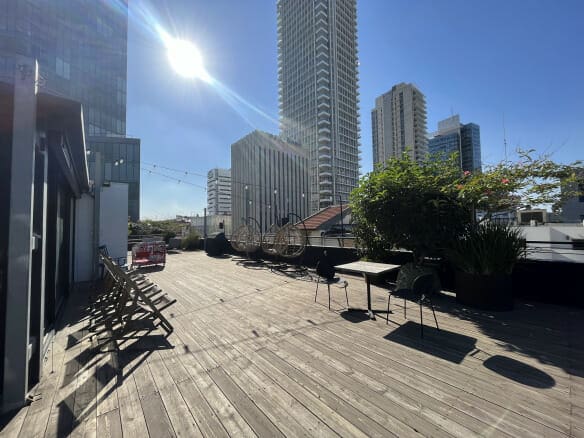 מרפסת גג שטופת שמש עם ריצוף עץ, כיסאות ושולחנות ריקים, עציצים ובניינים גבוהים בעיר ברקע.