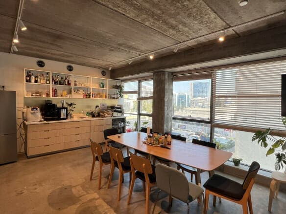 מטבח מודרני ופינת אוכל עם שולחן עץ, כיסאות מעורבים, חלונות גדולים עם תריסים, מדפים פתוחים, מקרר ונוף לעיר מבחוץ.