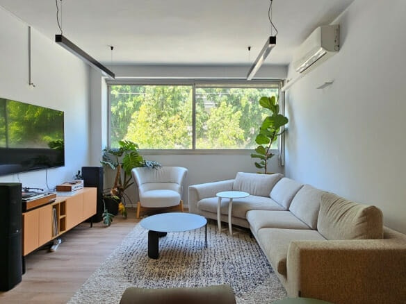 סלון מודרני עם ספה פינתית בצבע בז', כורסה לבנה, שולחנות עגולים, טלוויזיה עם מסך שטוח, יחידת מדיה מעץ, חלון גדול, צמחים ועיצוב מינימליסטי.