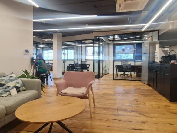 חלל משרדי מודרני עם אזור טרקלין, שולחן קפה עגול, כורסאות, מטבחון וחדרי ישיבות עם קירות זכוכית; אדם אחד עובד ליד שולחן ברקע.