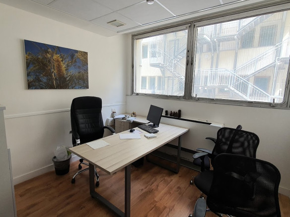 משרד קטן להשכרה ברחוב יגאל אלון, תל אביב, הכולל שולחן, שני כיסאות, מחשב, ניירות ועציץ. חלונות גדולים משקיפים על גרם מדרגות חיצוני; תמונה ממוסגרת מקשטת את הקיר.