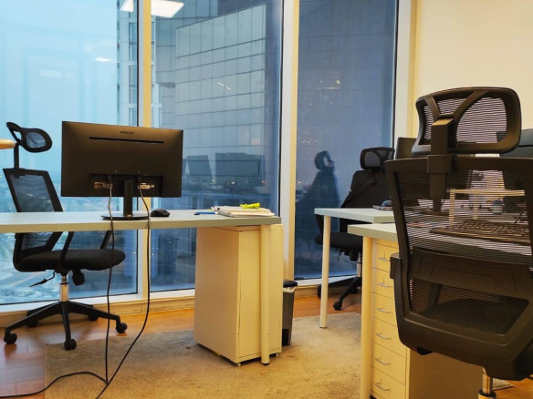 חלל משרדי מודרני עם שלושה כיסאות ארגונומיים, שני שולחנות, צג מחשב וחלונות גדולים המשקיפים על בניינים גבוהים בחוץ. הרצפה עשויה עץ, עם שטיח קטן מתחת לשולחנות.