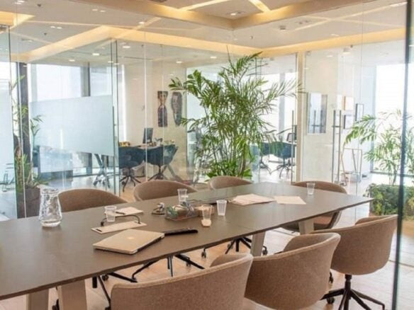 חדר ישיבות מודרני מסוג Plug&Play עם כיסאות בצבע בז' סביב שולחן גדול, ניירות ומחשבים ניידים על השולחן, צמחים ברקע וקירות זכוכית – מושלם לפגישה הבאה שלכם במשרד להשכרה בשרונה.