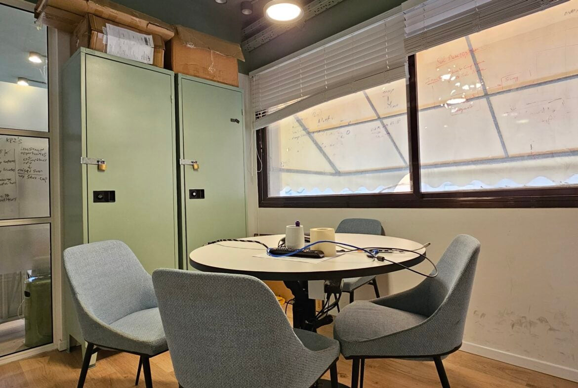 חדר ישיבות קטן עם ארבעה כיסאות סביב שולחן עגול, לוקרים ירוקים, לוח לבן עם פתקים וחלונות גדולים עם תריסים פתוחים חלקית במתחם משרדים להשכרה בורסה רמת גן.