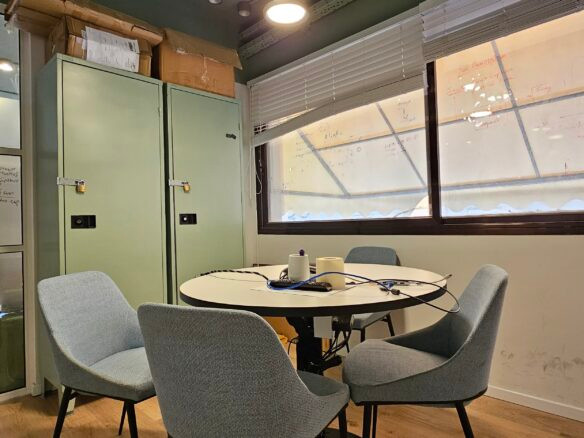 חדר ישיבות קטן עם ארבעה כיסאות סביב שולחן עגול, לוקרים ירוקים, לוח לבן עם פתקים וחלונות גדולים עם תריסים פתוחים חלקית במתחם משרדים להשכרה בורסה רמת גן.