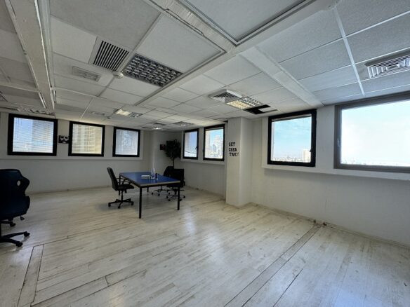 משרד מרווח להשכרה בשרונה, עם קירות ורצפה לבנים, שולחן כחול, שלושה כסאות וחלונות גדולים שמכניסים אור טבעי.