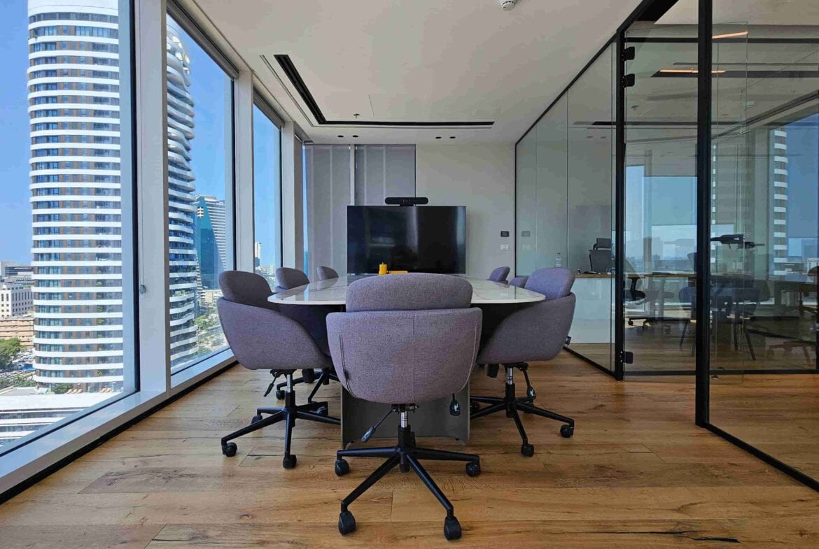 חדר ישיבות מודרני עם שולחן עגול, שישה כיסאות אפורים, חלונות גדולים המשקיפים על בנייני העיר במגדל SKY, וקירות זכוכית המפרידים בין המשרד במגדל לשאר חלל המשרדים.