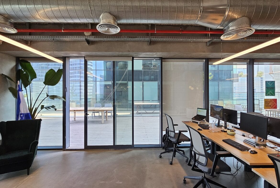 משרד יפה ומודרני עם שולחנות, מחשבים וכסאות הפונים לדלתות זכוכית גדולות הנפתחות למרפסת פרטית המשקיפה על בנייני העיר במגדלי הארבעה.
