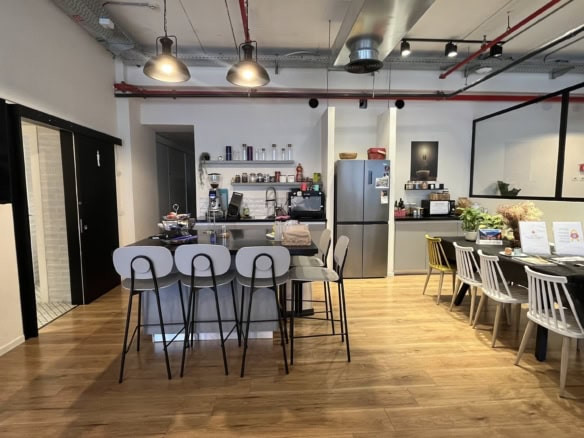 מטבח משרדי מודרני במשרד מעוצב במגדלי אלון, הכולל פינת קפה, כיסאות בר גבוהים ליד הדלפק, שולחן אוכל עם כיסאות, ומגוון מכשירים וציוד על מדפים ודלפקים. אידיאלי להשכרה.