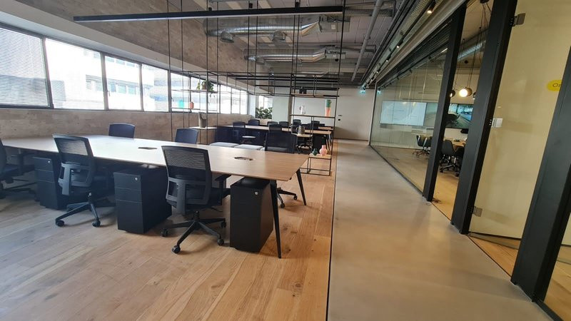 משרד מודרני בעל חלל פתוח עם רצפות עץ, שולחנות ארוכים, כיסאות משרדיים שחורים, חלונות גדולים וחדרי ישיבות עם קירות זכוכית. מושלם להשכרת עמדות עבודה ליד רכבת השלום. החלל ריק ומלא באור טבעי.