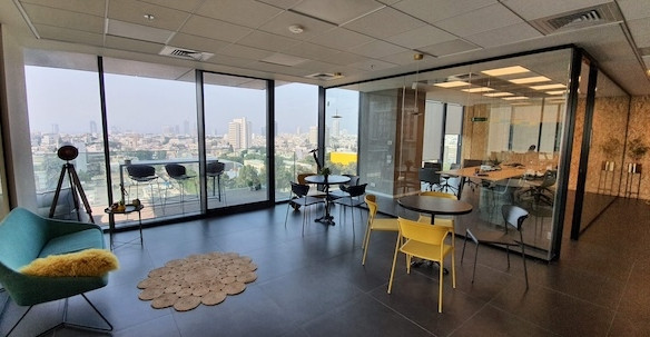 טרקלין מודרני ומעוצב עם קירות זכוכית, שולחנות עגולים, כיסאות, ספה ונוף פנורמי של העיר במגדלי הארבעה; חדר ישיבות סמוך נראה מבעד למחיצת הזכוכית.