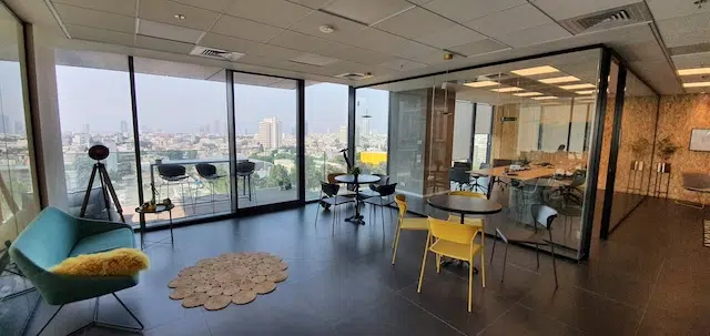 טרקלין מודרני ומעוצב עם קירות זכוכית, שולחנות עגולים, כיסאות, ספה ונוף פנורמי של העיר במגדלי הארבעה; חדר ישיבות סמוך נראה מבעד למחיצת הזכוכית.