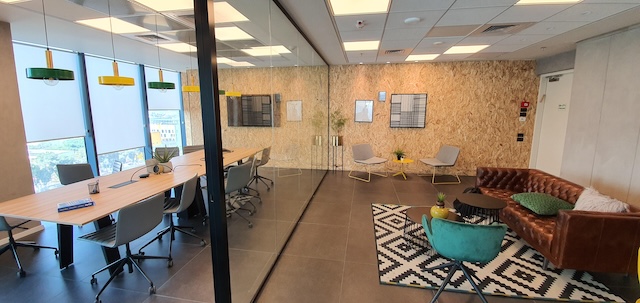 משרד מעוצב עם אווירה ייחודית, עם שולחן ישיבות בצד שמאל ואזור ישיבה עם כיסאות, ספה וקישוטים בצד ימין, המופרדים על ידי קיר זכוכית.