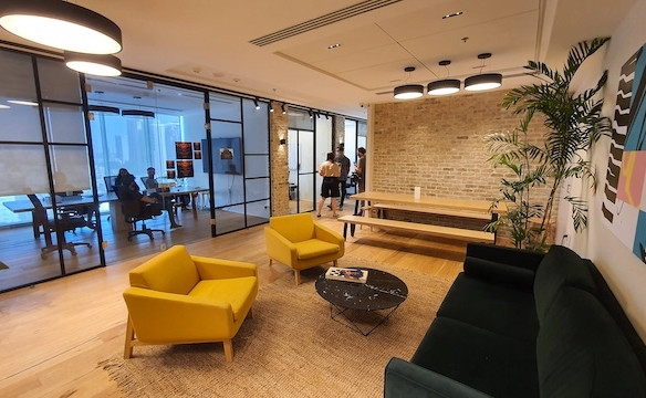 טרקלין משרדי מודרני עם כיסאות צהובים, ספה שחורה ושולחן קפה, הממוקם במגדל WE תל אביב. ברקע ניתן לראות חדר ישיבות עם אנשים בישיבה – מושלם להשכרת משרדים.
