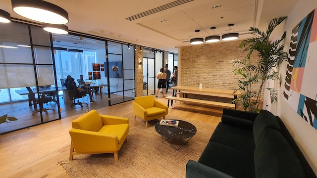 טרקלין משרדי מודרני עם כיסאות צהובים, ספה שחורה ושולחן קפה, הממוקם במגדל WE תל אביב. ברקע ניתן לראות חדר ישיבות עם אנשים בישיבה – מושלם להשכרת משרדים.