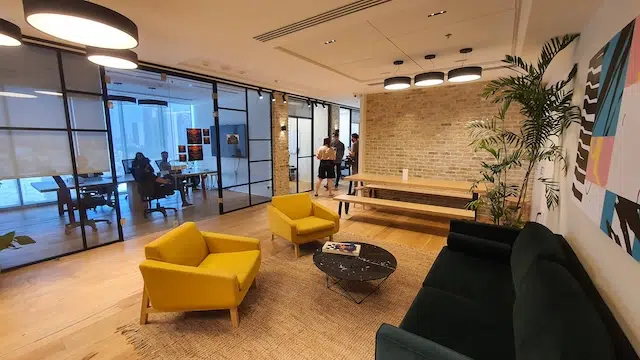 טרקלין משרדי מודרני עם כיסאות צהובים, ספה שחורה ושולחן קפה, הממוקם במגדל WE תל אביב. ברקע ניתן לראות חדר ישיבות עם אנשים בישיבה – מושלם להשכרת משרדים.