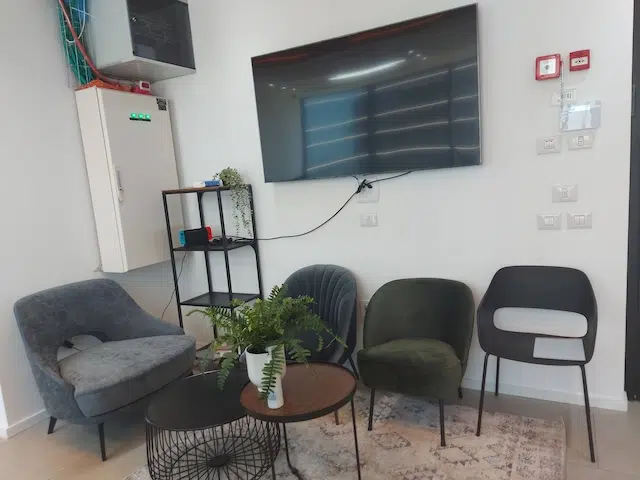 אזור המתנה במגדלי חג'ג' שרונה עם ארבעה כיסאות, שולחן קפה עגול עם עציצים, טלוויזיה המותקנת על הקיר ומדף קטן – מושלם לצרכי משרד להשכרה או משרד 100 מ