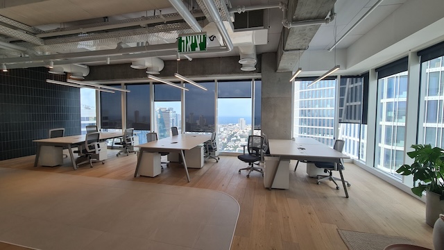 משרד מודרני בעל חלל פתוח עם מספר שולחנות וכסאות ריקים, חלונות גדולים עם נוף לעיר בקומה מרוהטת בתל אביב, רצפות עץ וצינורות חשופים בתקרה.