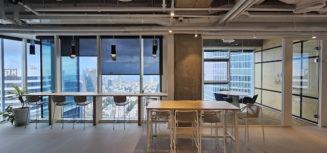 חלל משרדי מודרני עם שולחנות גבוהים, כיסאות, חלונות גדולים המשקיפים על העיר, צינורות תקרה חשופים ומרפסת להשכרה בקומה עצמאית במגדל סקיי תל אביב.