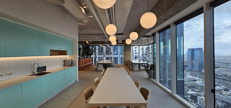 מטבח ומטבח מודרני עם ארונות מנטה, שולחנות ארוכים, תאורה עגולה בתקרה, חלונות גדולים ונוף של קו הרקיע העירוני במגדל סקיי ת״א; תיהנו מקומה מרוהטת במלואה עם גישה למרפסת להשכרה.