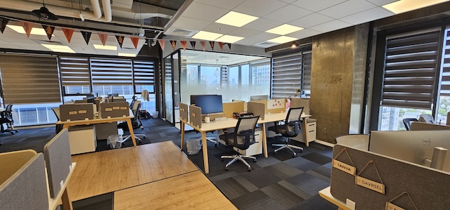 שטח משרדי מודרני להשכרה במגדלי אלון, תל אביב, הכולל מספר שולחנות, כיסאות, מחשבים, חלונות גדולים ודגלים דקורטיביים התלויים מהתקרה.