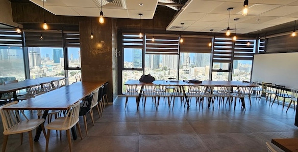 חדר ישיבות מודרני ומרווח למשרד להשכרה במגדלי אלון, תל אביב, עם חלונות גדולים, שולחנות וכסאות מעץ, תאורה תלויה ונוף עוצר נשימה של העיר ברקע.