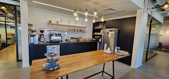 מטבח משרדי מודרני במשרד להשכרה במגדלי אלון, תל אביב, הכולל שולחן עץ ארוך, מכונות קפה, חטיפים על הדלפק ומדפים עם ספלים ואספקה על רקע אריחי קיר לבנים.