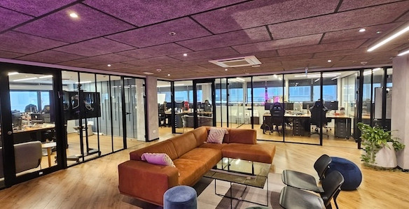 חלל משרדים מודרני בבית יכין שרונה, הכולל חדרי ישיבות עם קירות זכוכית, ספה חומה, כיסאות, ריצוף עץ ואנשים העובדים ליד שולחנות ברקע. תיהנו מהאווירה המרווחת של משרד גדול.