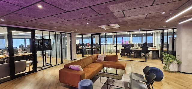 חלל משרדים מודרני בבית יכין שרונה, הכולל חדרי ישיבות עם קירות זכוכית, ספה חומה, כיסאות, ריצוף עץ ואנשים העובדים ליד שולחנות ברקע. תיהנו מהאווירה המרווחת של משרד גדול.